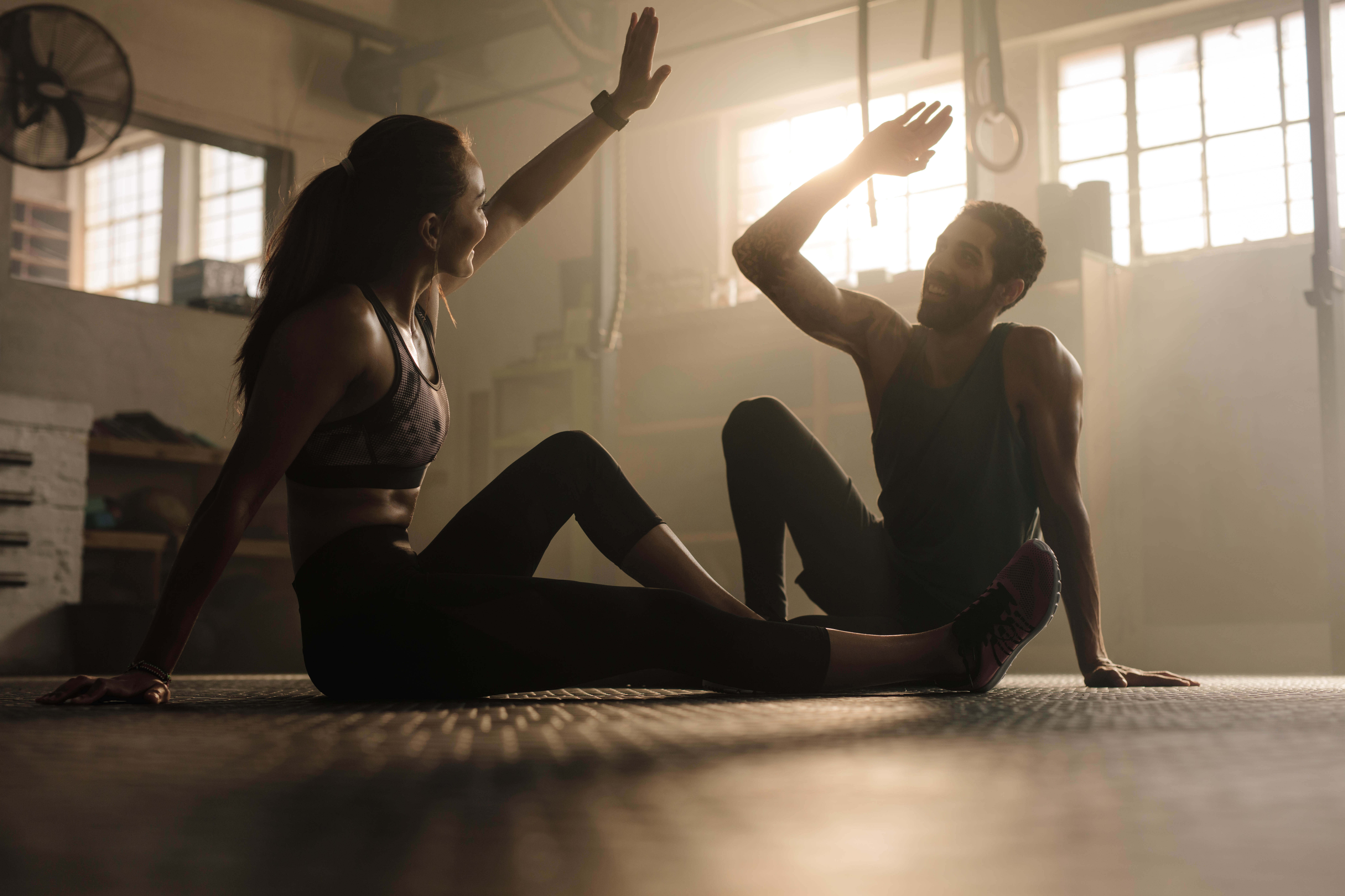 Young, fit couple sitting on the floor of a gym, hands raised as if they're about to high five